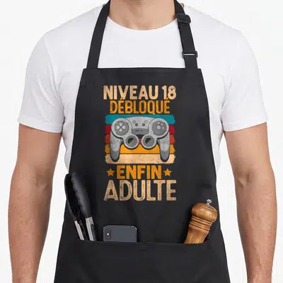 Tablier de cuisine noir avec illustration de manette de jeu et texte Niveau 18 Débloqué Enfin Adulte sur fond coloré vintage.