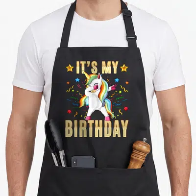 Tablier de cuisine noir avec une licorne blanche faisant le dab, des étoiles colorées et le texte It's My anniversaire en lettres dorées.