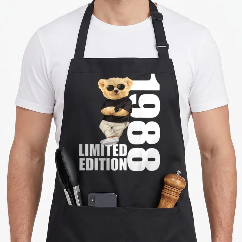 Tablier de cuisine noir avec ours aux lunettes de soleil, bras croisés et texte 1988 Limited Edition imprimé en blanc.