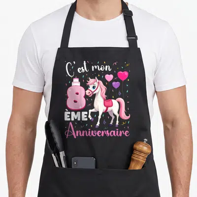 Tablier de cuisine noir avec illustration d'un poney blanc aux cheveux roses, un gâteau et des ballons colorés.