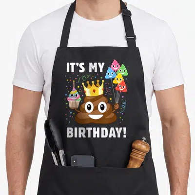 Tablier de cuisine noir avec illustration d'un personnage marron souriant portant une couronne dorée et tenant des ballons festifs.
