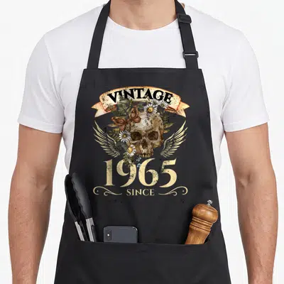 Tablier de cuisine noir avec une tête de mort vintage, des ailes blanches, des marguerites et le texte Vintage 1965 Since imprimé en centre.