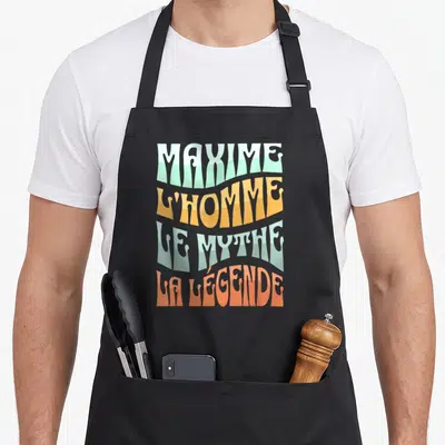 Tablier de cuisine noir avec typographie rétro colorée proclamant Maxime l'homme le mythe et la légende sur fond uni.