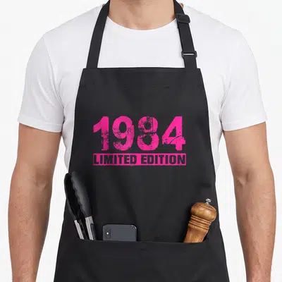 Tablier de cuisine noir avec l'inscription 1984 Limited Edition en rose vif et texture vieillie au centre.