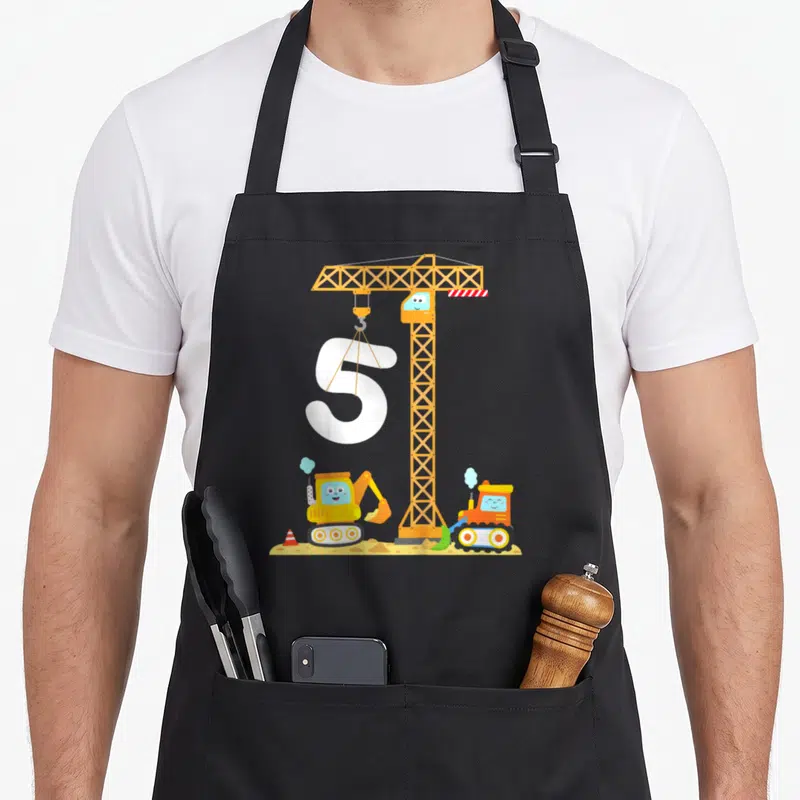 Tablier de cuisine noir avec illustration de grue, pelleteuse, bulldozer et le chiffre 5 blanc sur fond de chantier.