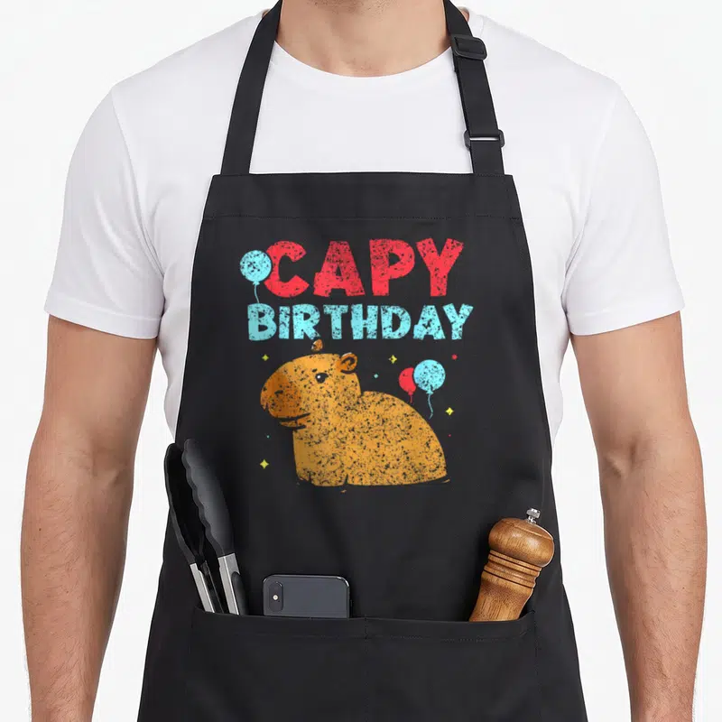 Tablier de cuisine noir avec illustration d'un capybara orange et le texte CAPY anniversaire entouré de ballons colorés.