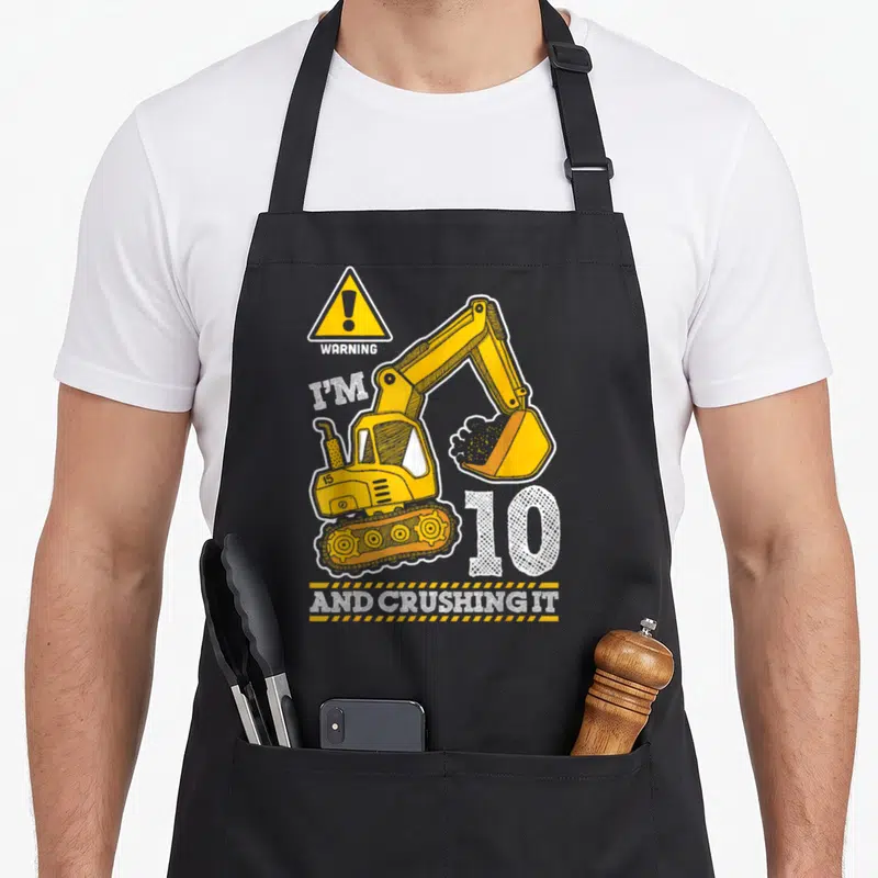 Tablier de cuisine noir avec pelleteuse jaune, panneau warning et texte I'm 10 and crushing it imprimé en blanc et orange.