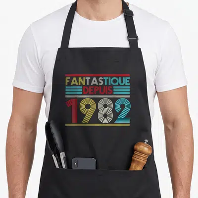Tablier de cuisine noir avec inscription « Fantastique Depuis 1982 » en lettres rayées multicolores style années eighty.