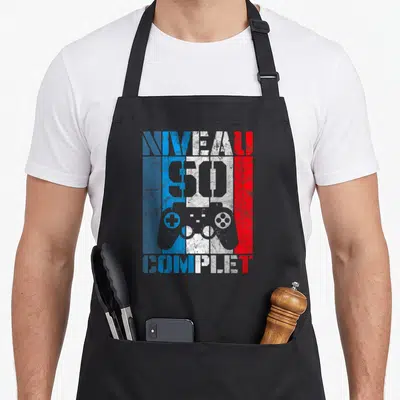 Tablier de cuisine noir avec inscription Niveau complet 50 ans et manette de jeu vidéo colorée sur fond texturé.