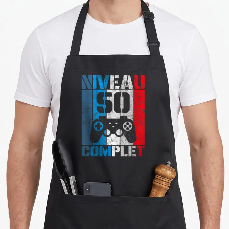 Tablier de cuisine noir avec inscription Niveau complet 50 ans et manette de jeu vidéo colorée sur fond texturé.