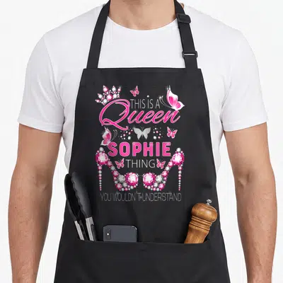 Tablier de cuisine noir avec inscription Reine Sophie Thing entourée de talons roses et papillons scintillants.