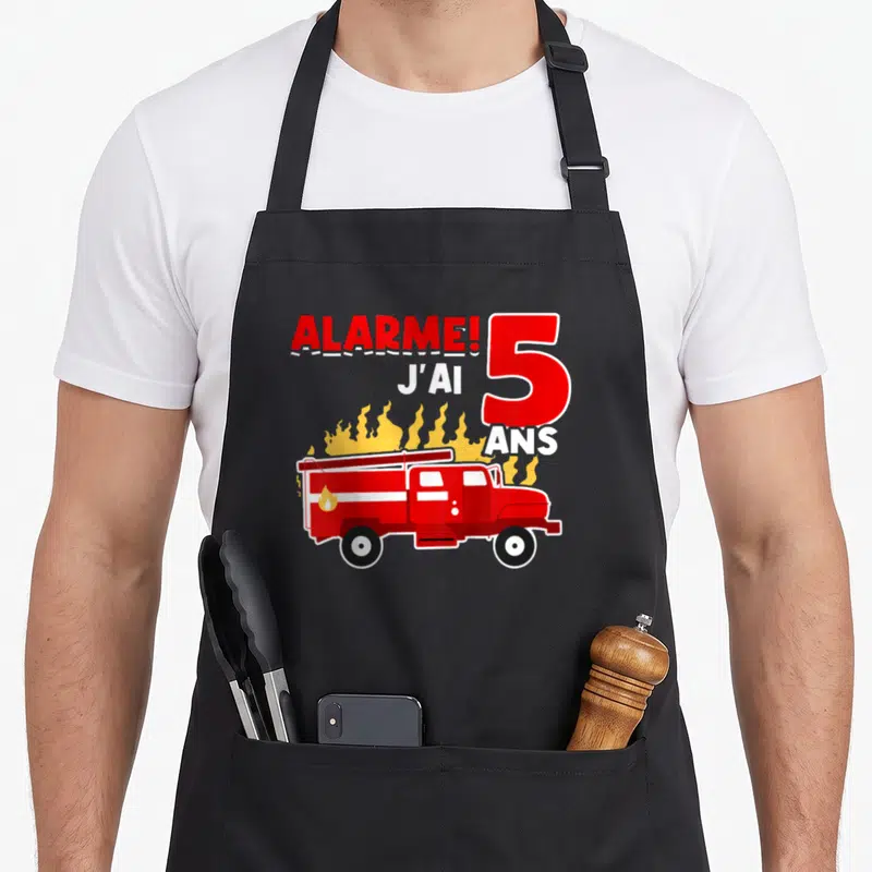 Tablier de cuisine noir avec illustration de camion de pompiers rouge et texte alarme j'ai 5 ans sur fond de flammes jaunes.