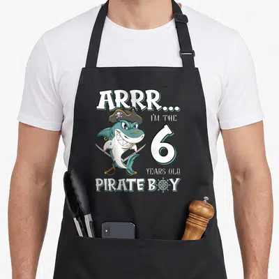 Tablier de cuisine noir avec un requin pirate tenant des sabres et le chiffre 6 en grosses lettres blanches.