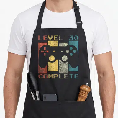 Tablier de cuisine noir avec une illustration de manette de jeu vidéo Level 30 Complete en style vintage multicolore.