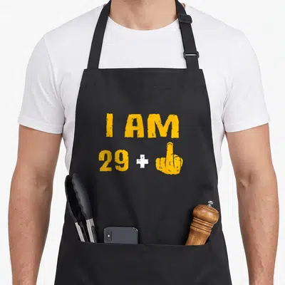 Tablier de cuisine noir avec l'inscription jaune I AM 29 et un geste d'insultation blanc.
