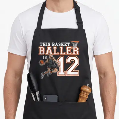Tablier de cuisine noir avec illustration d'un joueur de basketball et le texte This Basketballer Is 12 en blanc et orange.