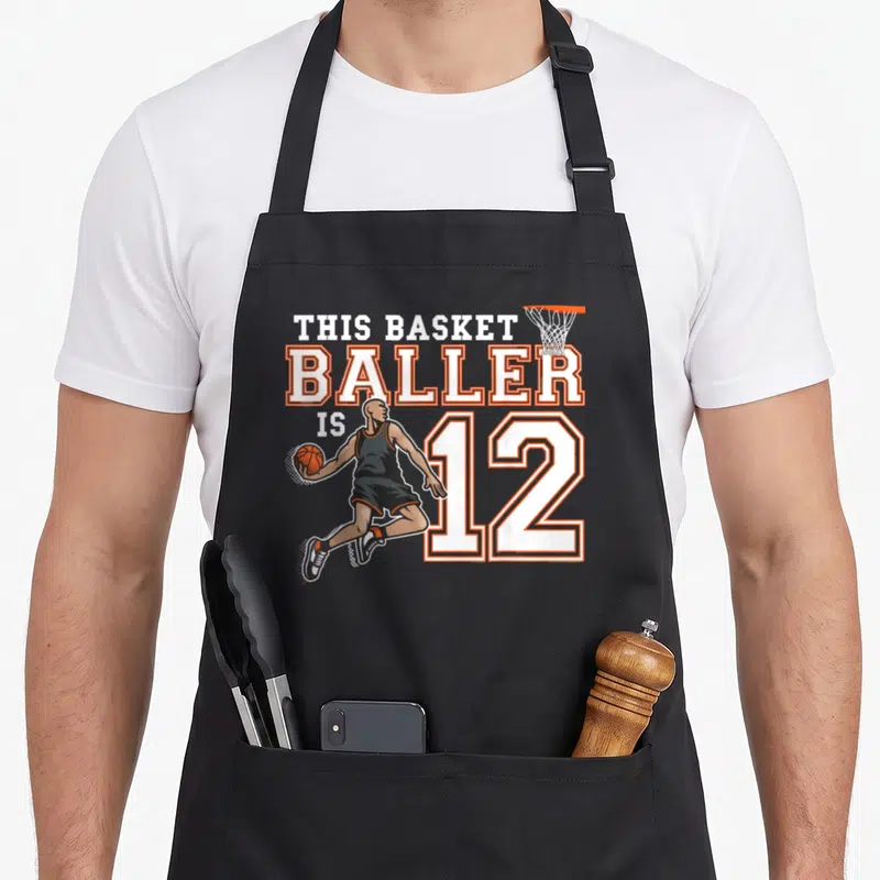 Tablier de cuisine noir avec illustration d'un joueur de basketball et le texte This Basketballer Is 12 en blanc et orange.