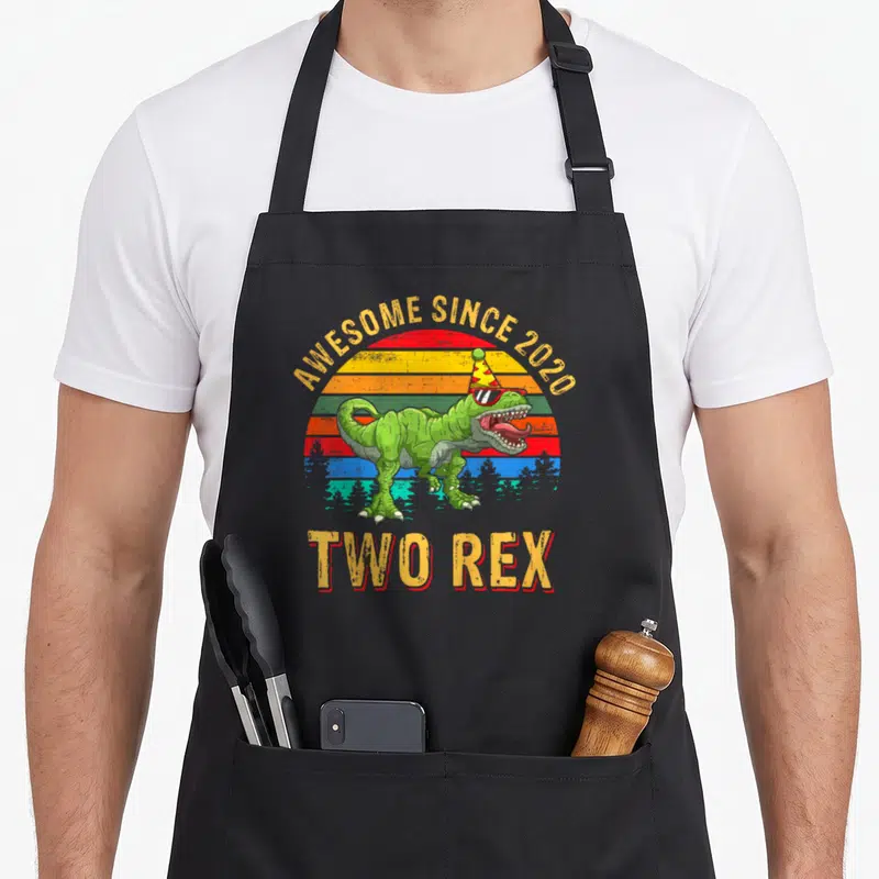 Tablier de cuisine noir avec un dinosaure vert portant chapeau et lunettes devant un soleil arc-en-ciel.