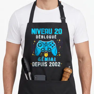 Tablier de cuisine noir avec illustration manette bleue et texte Niveau 20 Débloqué Génial Depuis 2002 entouré d'étoiles.