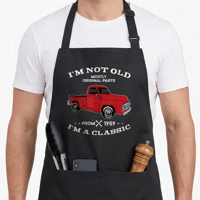 Tablier de cuisine noir avec illustration de camion rouge vintage et texte blanc humoristique sur fond uni.