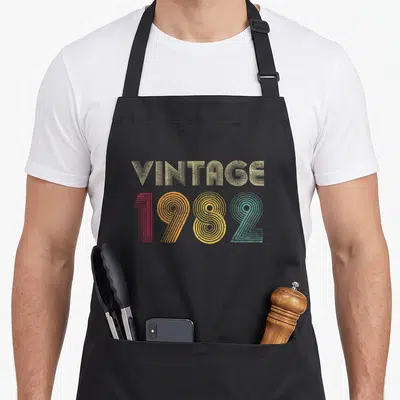 Tablier de cuisine noir avec linscription vintage 1982 en lettres colorées et motif rayé rétro.