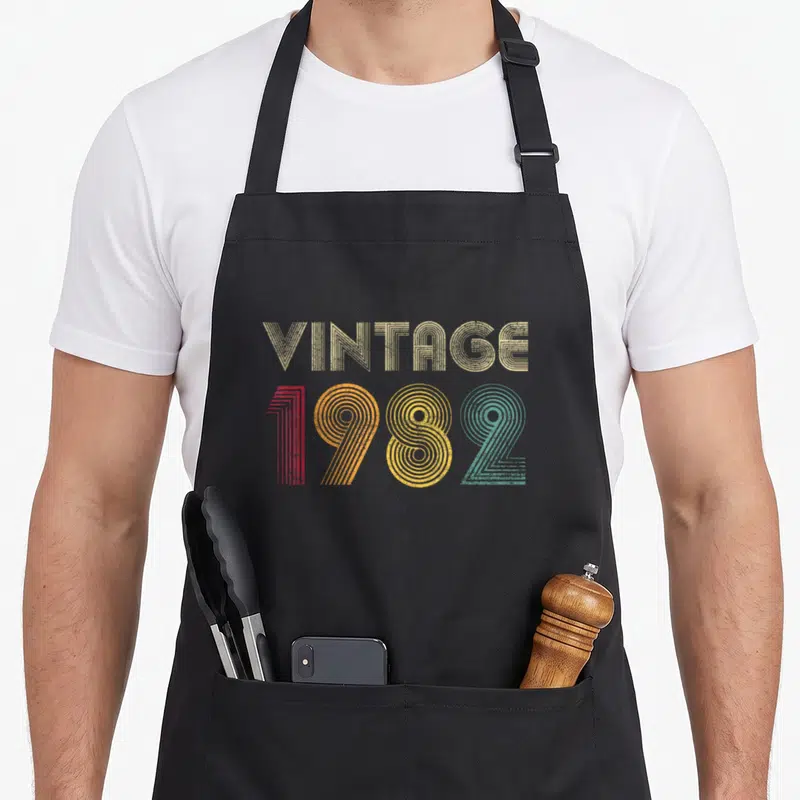 Tablier de cuisine noir avec l'inscription vintage 1982 en lettres colorées et motif rayé rétro.