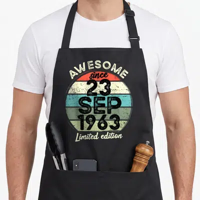 Tablier de cuisine noir avec motif circulaire coloré affichant « Awesome since 23 SEP 1963 Limited edition » en typographie rétro.
