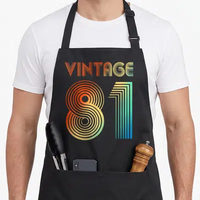 Tablier de cuisine noir avec l'inscription Vintage 1981 en lettres colorées au style graphique des années quatre-vingt.