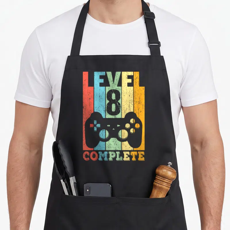 Tablier de cuisine noir avec l'inscription Level 8 Complete et une manette de jeu multicolore sur fond de barres verticales.