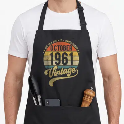 Tablier de cuisine noir avec un motif circulaire retro affichant 1961 et le mot Vintage en lettres dorées sur fond de soleil couchant.