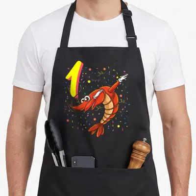 Tablier de cuisine noir avec une crevette rose animée, le chiffre 1 jaune et des confettis multicolores.