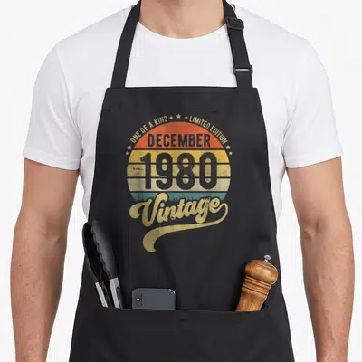 Tablier de cuisine noir avec motif circulaire rétro affichant December 1980 et Vintage en lettres dorées et grises.