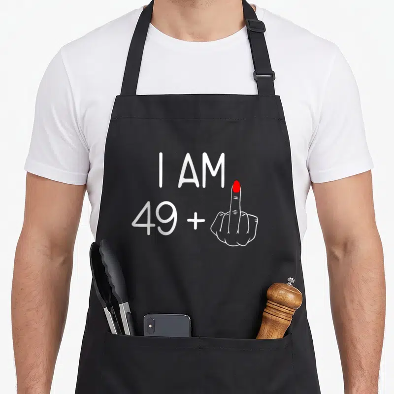 Tablier de cuisine noir avec inscription blanche I Am 49 Plus 1 et main au majeur à ongle rouge.
