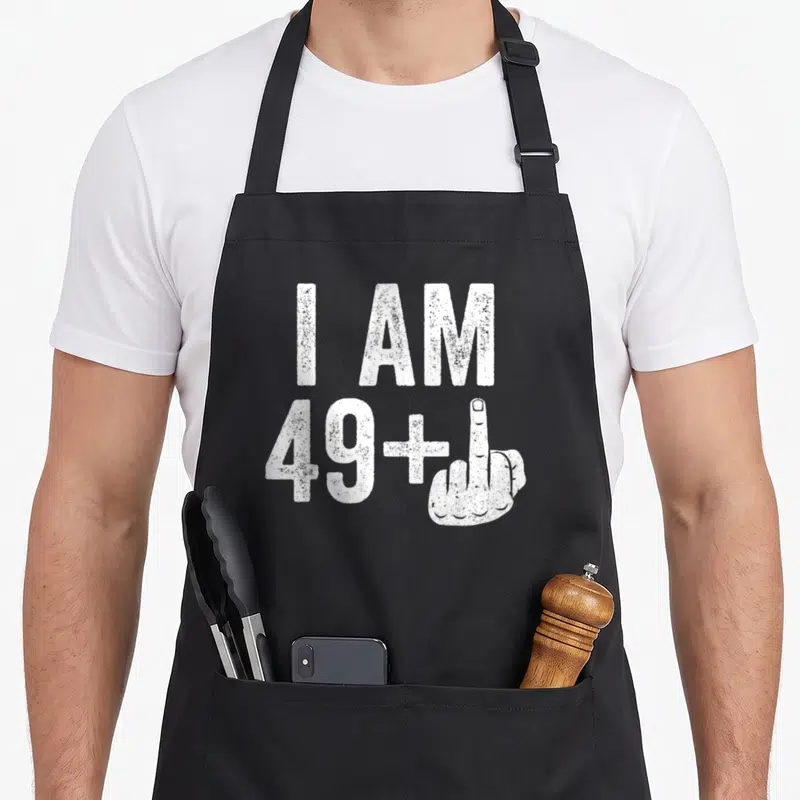 Tablier de cuisine noir avec inscription blanche I AM 49+ et illustration de main levée.
