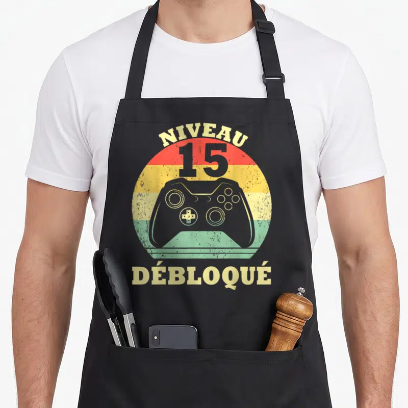 Tablier de cuisine noir avec illustration manette de jeu et texte Niveau 15 Débloqué sur fond arc-en-ciel rétro.