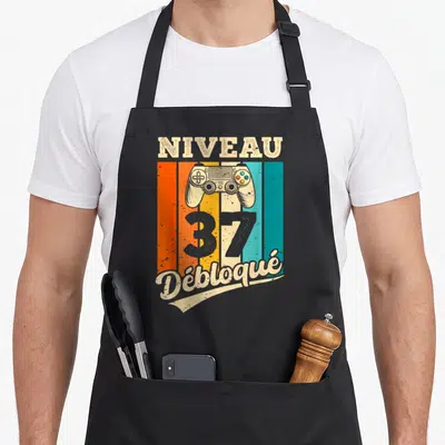 Tablier de cuisine noir avec manette de jeu et texte Niveau 37 Débloqué sur fond coloré vintage.