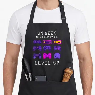 Tablier de cuisine noir avec manettes de jeux vidéo violettes et texte Un geek ne vieillit pas il level-up.