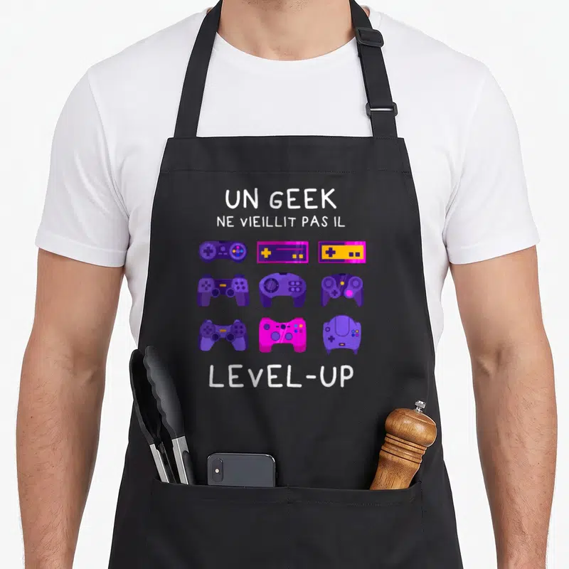 Tablier de cuisine noir avec manettes de jeux vidéo violettes et texte Un geek ne vieillit pas il level-up.