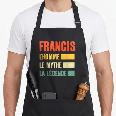 Tablier de cuisine noir avec le texte Francis L'Homme Le Mythe La Légende en lettres orange jaune crème et vert menthe.