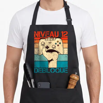 Tablier de cuisine noir avec main tenant une manette et texte Niveau 12 Débloqué sur fond rayé vintage.
