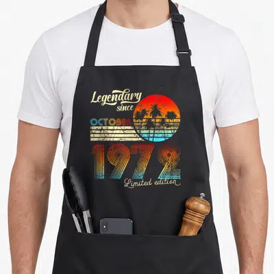 Tablier de cuisine noir avec illustration rétro « Legendary since October 1972 Limited edition » et soleil aux palmiers colorés.