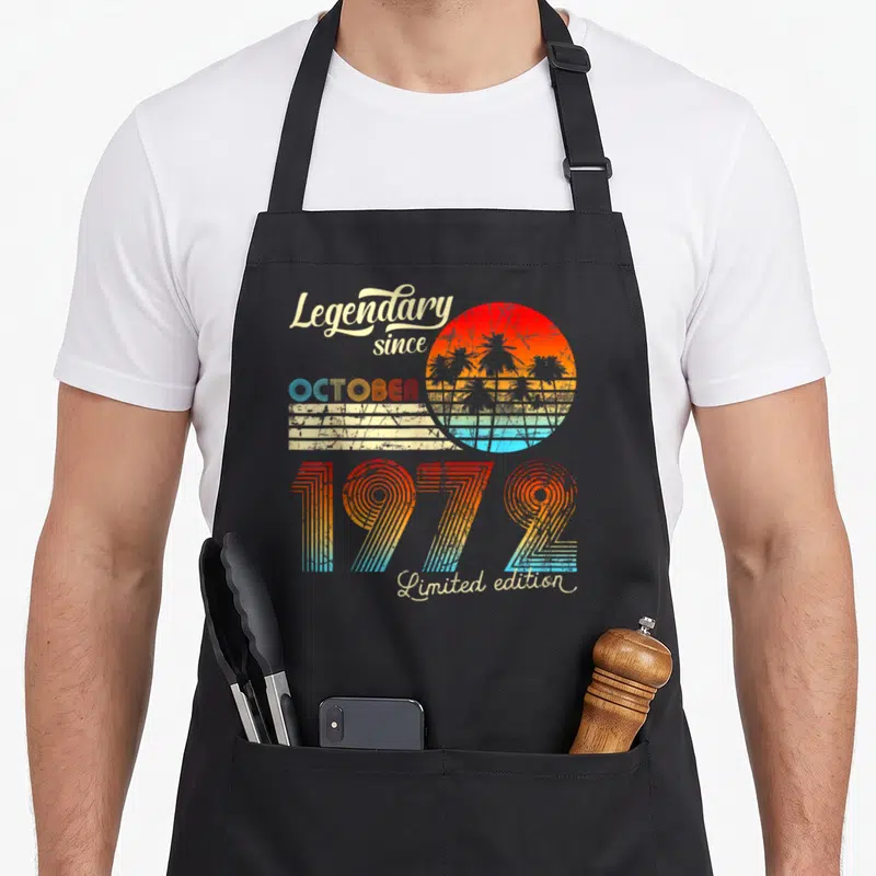 Tablier de cuisine noir avec illustration rétro « Legendary since October 1972 Limited edition » et soleil aux palmiers colorés.