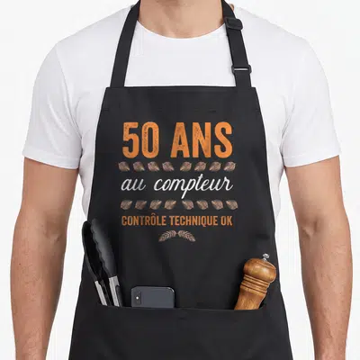 Tablier de cuisine noir avec inscription 50 ans au compteur contrôle technique ok et motifs de feuilles oranges.