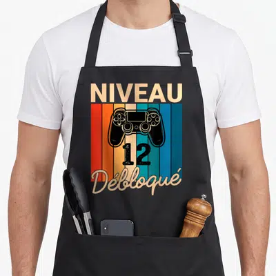 Tablier de cuisine noir avec manette de jeu et texte Niveau 12 Débloqué sur fond rayé coloré.