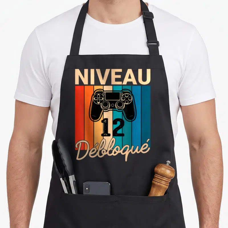 Tablier de cuisine noir avec manette de jeu et texte Niveau 12 Débloqué sur fond rayé coloré.