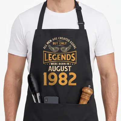 Tablier de cuisine noir avec inscription dorée Les légendes d'anniversaire sont nées en août 1982 surmontée de deux ailes stylisées.