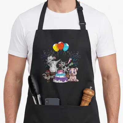Tablier de cuisine noir avec des animaux de la ferme célébrant un anniversaire autour d'un gâteau et de ballons colorés.
