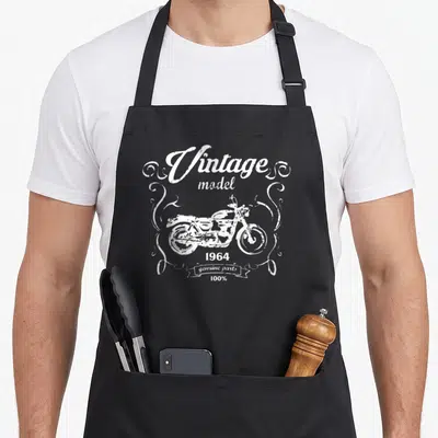Tablier de cuisine noir avec illustration blanche de moto vintage et texte model 1964 genuine parts entouré de volutes décoratives.