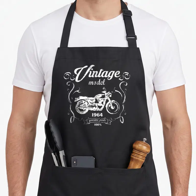 Tablier de cuisine noir avec illustration blanche de moto vintage et texte model 1964 genuine parts entouré de volutes décoratives.