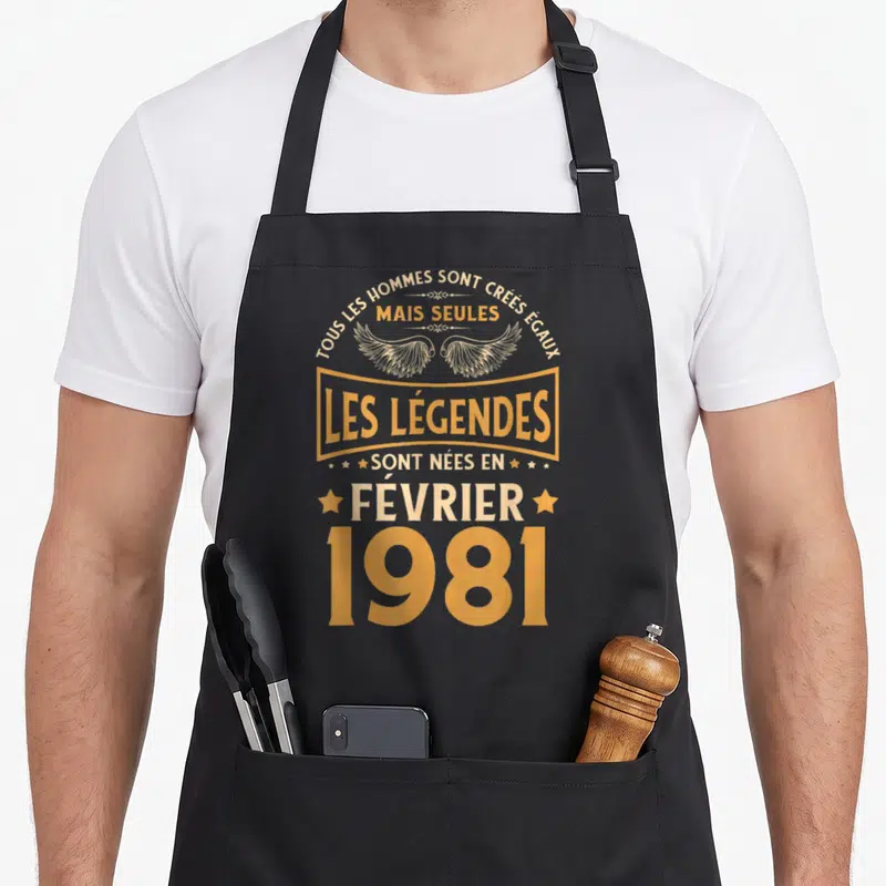 Tablier de cuisine noir avec inscription dorée Les Légendes Sont Nées en Février 1981 orné d'ailes stylisées.